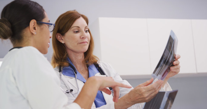 Two Medical Colleagues Looking At Xrays Of Patients Wrist And Hand Inside Office. Attractive Hispanic Doctor Discussing X Rays Of Hands With Older Colleague