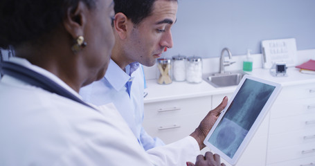 Fototapeta na wymiar Middle aged black doctor using electronic tablet to view patient x ray of spine and pelvis. Young latino patient looking at x ray of his body on electronic notebook tablet