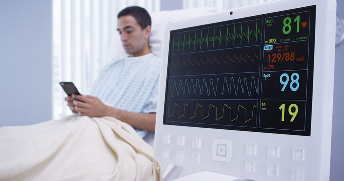 Tight Shot Of EKG Monitor And Male Patient Lying Down On Hospital Bed Texting On Phone. Close Up Of Patients Vitals Reading On Monitor Screen While He Reads Text Messages On Smartphone