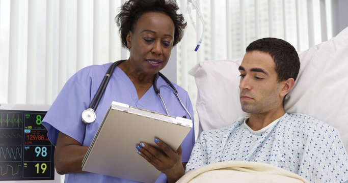 Middle Aged African Nurse Taking Notes Of Male Patients Current Health Condition. Senior Female RN Recording Patients Condition Post Surgery