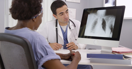 Old African patient paying doctor visit to review results from x-rays of lungs. Young Hispanic...