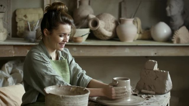 PAN of happy young woman in apron smiling and throwing vase on spinning pottery wheel in workshop