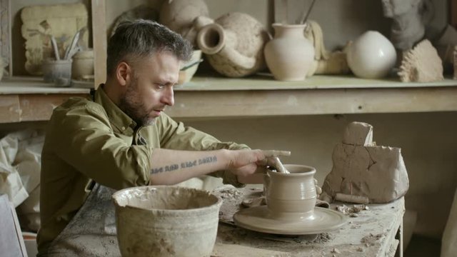 PAN of focused bearded male artisan in apron wetting his hands and throwing vase on spinning pottery wheel in workshop
