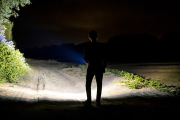 back view of man standing outdoor on night forest road lighting the way with electric flashlight d - obrazy, fototapety, plakaty