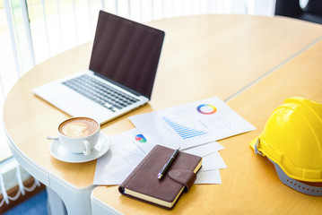 Workplace Desktop table with Laptop , Document Papers , Coffee Cup , Smartphone and Safety Engineer Helmet , Modern Office room with white curtains in sunny day