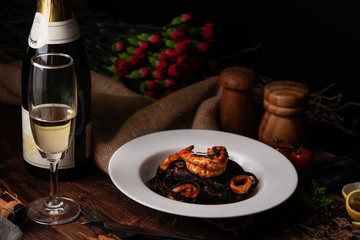 Black fettuccine spaghetti pasta with shrimp and squid topping with green parsley in white dish on woodden table with white wine and flowers, selective focus