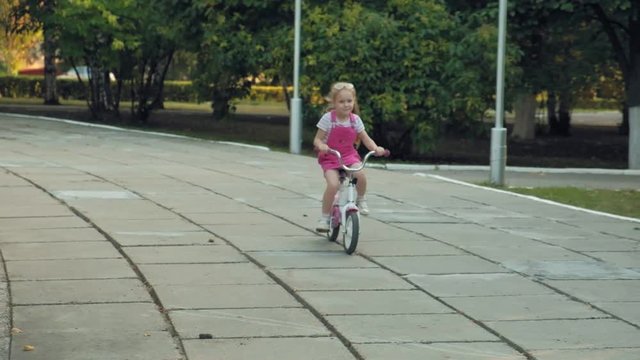 A happy, beautiful, little girl with long blond hair in a pink skirt and jumper rides a children's bike on the road, she smiles. Super slow motion