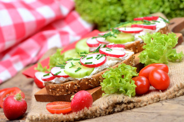 Wholemeal grain bread with curd cheese, fresh radish, cucumber and tomatoes on a wooden cutting board - concept of healthy fitness breakfast or snack