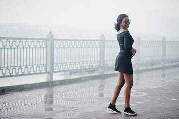 Fototapeta na wymiar African american girl in sunglasses, black clothes and shirt posed outdoor. Fashionable black woman against rain of fountains.