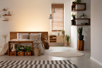 Real photo of a warm bedroom interior with wooden boxes and shelves, double bed and plant. Empty wall, place your logo