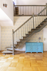 Real photo of a simple corridor with gray, wooden stairs, blue cupboards and floral wallpaper in...