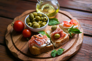 Appetizing bruschettes with jamon, tomatoes, basil, mozzarella, olives and oil on a dark wooden board