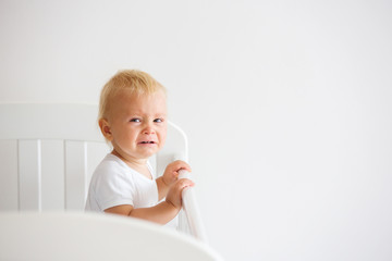 Portrait of bored baby standing in crib. Sad little baby, crying in bed