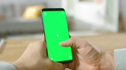 Close-up of a Man Uses Green Mock-up Screen Smartphone and Does Touchscreen Gestures. Touching Mobile Phone Chroma Key Screen. In the Background Cozy Homely Atmosphere.