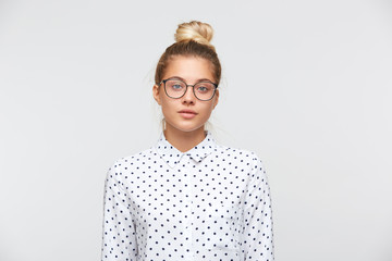 Closeup of serious beautiful young woman with bun wears polka dot shirt and glasses feels unhappy...