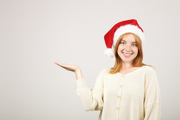 Close up portrait of beautiful redheaded young woman wearing Santa Claus hat & white sweater with satisfied facial expression. Cute female celebrating New Year. Copy space, isolated on gray background