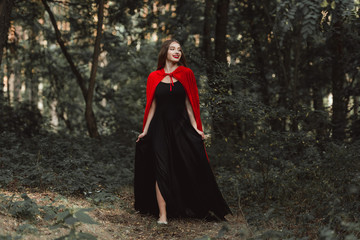 beautiful mystic girl in black dress and red cloak walking in forest
