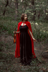 elegant mystic girl in black dress and red cloak holding violin in dark woods