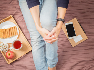 Flatlay of woman in jeans sitting on her bed hugging her knees, with smartphone, notepad, breakfast with tea, top view