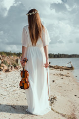 back view of girl in elegant white dress holding violin on seashore © LIGHTFIELD STUDIOS