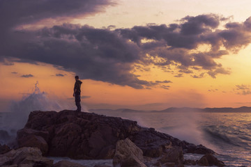 A man stands on a rock and looks at the raging sea, waves and splashes. Sunset on the sea after the storm. Man and the sea.