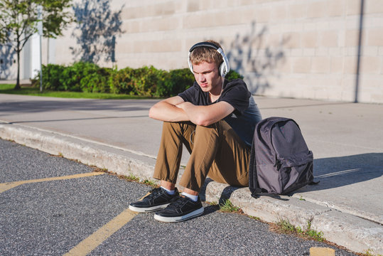 Sad teenage boy sitting on curb near school while listening to music on his headphones.