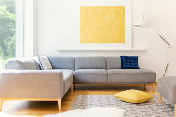 A minimalist, yellow poster and a white, industrial floor lamp in a sunny living room interior with a patterned rug and vibrant decorations