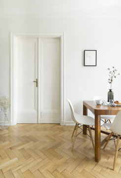 A bright dining room interior with a wooden table and chairs standing on a herringbone parquet against a white wall with double door. Real photo.