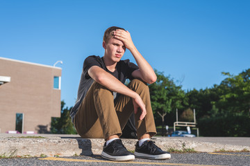 Upset teenager resting his head on his hand as he sits on the curb in front of his school.