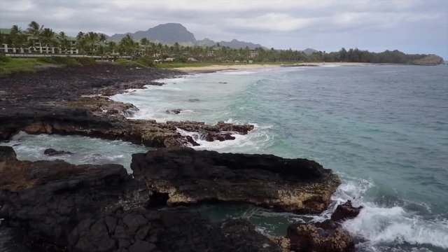 Bay with ocean waves, black lava rock, beach, palm trees, hotel and mountain