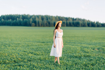Fototapeta premium Girl walking on the field, in a hat and summer dress. Smiling and laughing, beautiful sunset in the forest and in nature. Happy traveler, lifestyle.