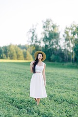 Fototapeta na wymiar Girl walking on the field, in a hat and summer dress. Smiling and laughing, beautiful sunset in the forest and in nature. Happy traveler, lifestyle.