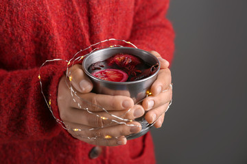 Woman holding metal cup of delicious mulled wine with Christmas garland on dark background, closeup