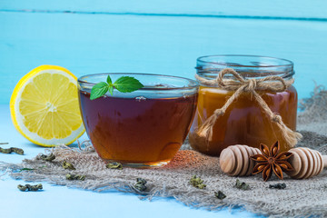 A cup of hot tea with a slice of fresh mint, a juicy, yellow lemon, a homemade honey jar and a honey spoons on a blue background. Tea drinking
