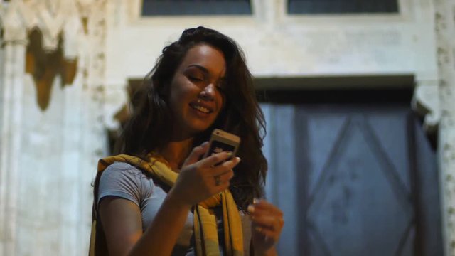 Joyful girl making selfie in evening. Young woman making selfie showing sign victory by fingers. Female tourist taking mobile selfie with v sign hand on old building background