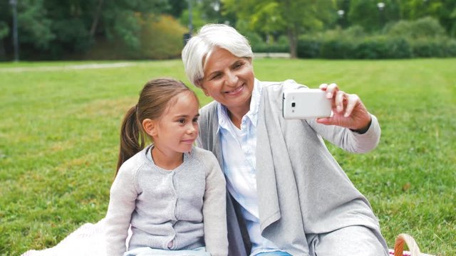 family, leisure and technology concept - happy grandmother and granddaughter having picnic and taking selfie by smartphone at summer park