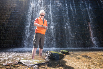 Young man with a blonde hair and beard posing for a camera in front of a waterfall in nature enviroment