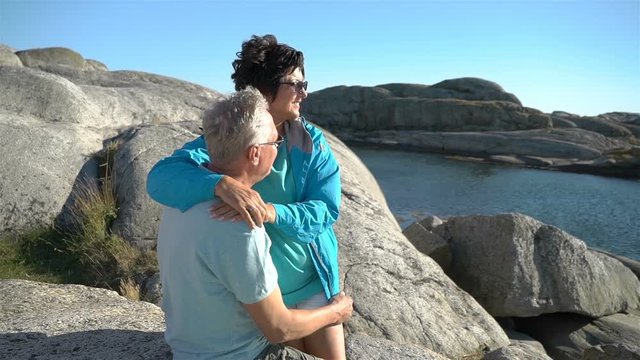 A happy loving mature couple enjoys a walk among the coastal stones on the seashore. They hold hands, admire the scenery, hug and kiss. Slow motion