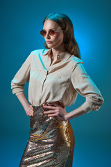 beautiful woman in fashionable golden glittering skirt and sunglasses posing with hands on waist isolated on blue