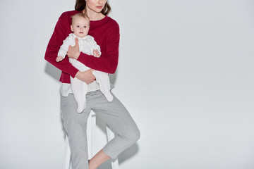 Obraz premium cropped shot of stylish young mother holding infant daughter and sitting on stool on grey