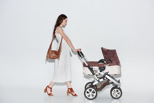 side view of woman in stylish white dress walking with baby stroller on grey