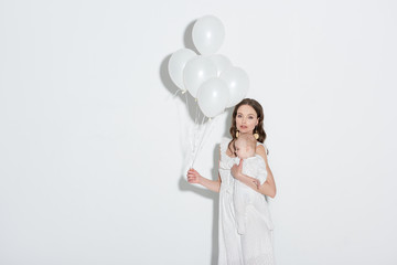 beautiful stylish young mother holding white balloons, carrying adorable infant daughter and looking at camera on grey