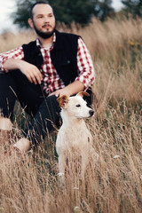 a male hipster sits in a park with a dog