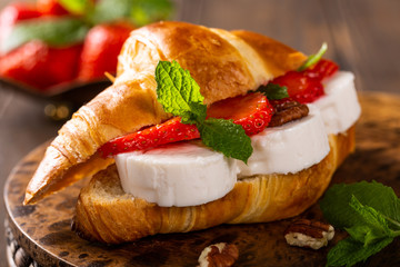 Fresh sandwich with croissant with goat cheese, strawberries, honey and pecans on wooden cutting board.