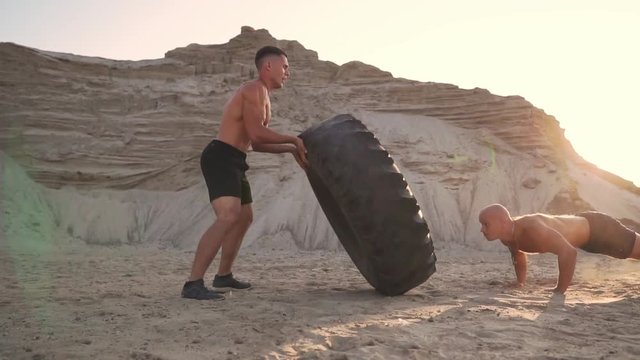 Two athletes train in active mode on the beach doing push-UPS and pushing a huge wheel