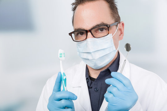 Ein Zahnarzt mit Mundschutz hält einen Zahnspiegel und eine Zahnbürste