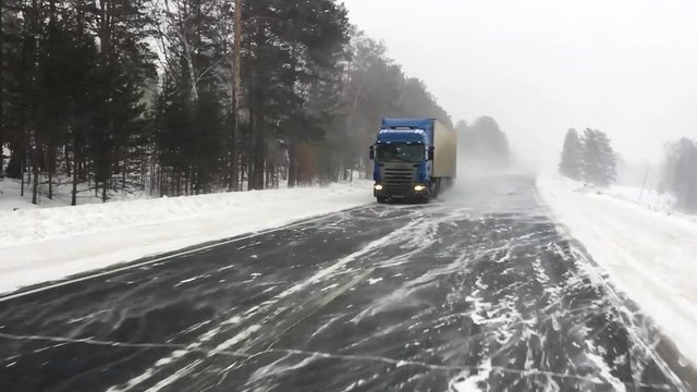  The car is on a snow-covered highway. Heavy truck with trailer carries cargo.  Winter road with a gusty wind and a snowstorm, slow motion. 