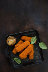 Cast-iron serving pan with roasted fish fingers, lemon and green basil. Flatlay on a dark brown metal surface with space, vertical shot