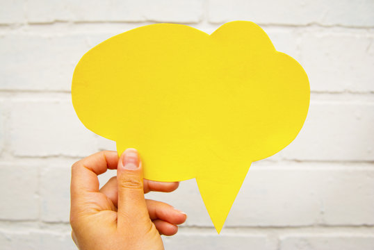 Hand holding an empty yellow hand-cuted paper speech bubble on white textured brick wall background. Isolated.