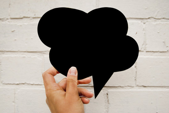Hand holding an empty black hand-cuted paper speech bubble on white brick wall textured background. Isolated.